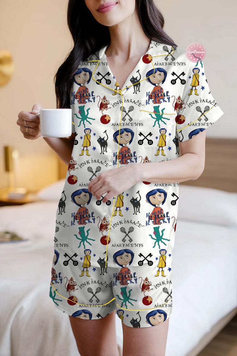 May include: White pajama set with a repeating pattern of cartoon characters, keys, and text. The set includes a short-sleeved top and shorts, trimmed with yellow piping. The pajama top has a button-down front. The person is holding a white mug.