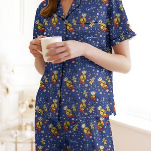 Peut inclure: Ensemble de pyjama bleu avec un motif r&eacute;p&eacute;t&eacute; d'un personnage de dessin anim&eacute;, d'&eacute;toiles et d'autres formes. Le haut du pyjama a un col en V et des manches courtes. Le mot "Coraline" est imprim&eacute; sur le haut et le bas. Une personne tient une tasse blanche.