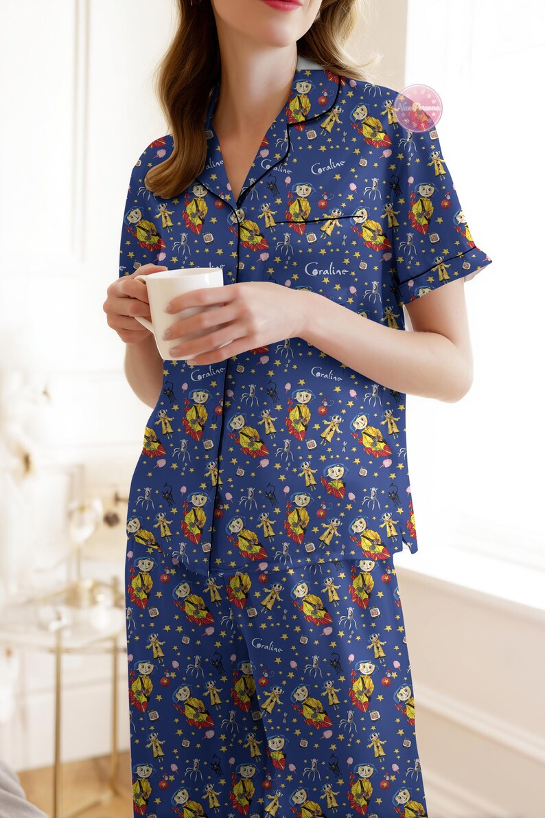 Peut inclure: Ensemble de pyjama bleu avec un motif r&eacute;p&eacute;t&eacute; d'un personnage de dessin anim&eacute;, d'&eacute;toiles et d'autres formes. Le haut du pyjama a un col en V et des manches courtes. Le mot "Coraline" est imprim&eacute; sur le haut et le bas. Une personne tient une tasse blanche.