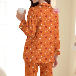 May include: Orange pajama set with a button and polka dot pattern. The long-sleeved top and pants feature a repeating design of white and red circles, and white buttons. The pajamas have black trim.