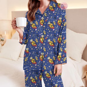 Peut inclure: Ensemble de pyjama bleu avec un motif r&eacute;p&eacute;t&eacute; de personnages de dessins anim&eacute;s, d'&eacute;toiles et le nom "Coraline". Le haut &agrave; manches longues et le pantalon sont en mati&egrave;re douce. La personne sur l'image tient une tasse blanche.