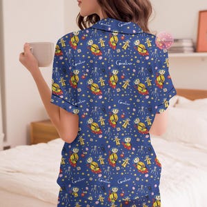 Peut inclure: Ensemble de pyjama bleu avec un motif r&eacute;p&eacute;t&eacute; d'un personnage de dessin anim&eacute;, d'&eacute;toiles et du mot "Coraline". Le haut &agrave; manches courtes et le short ont une bordure noire. La personne sur l'image tient une tasse beige.