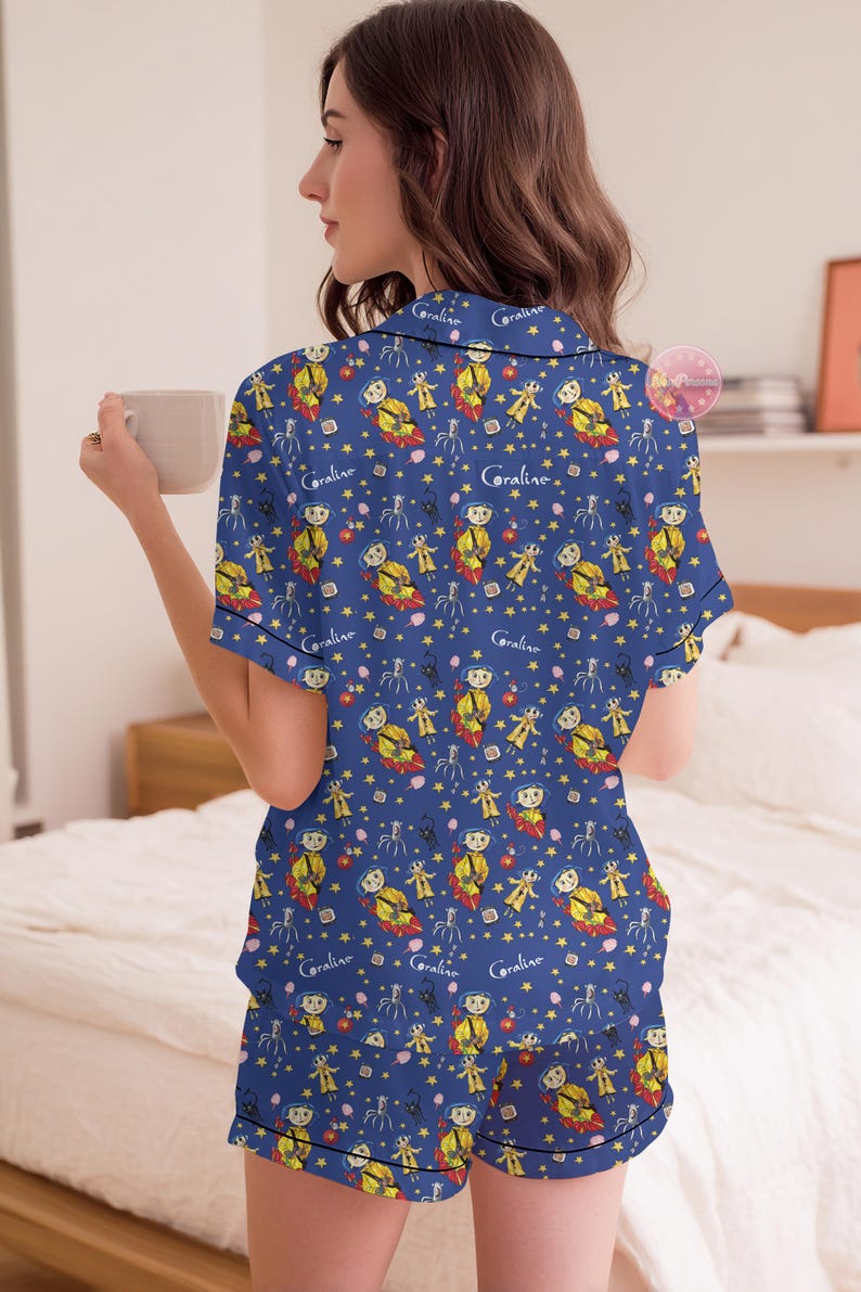 Peut inclure: Ensemble de pyjama bleu avec un motif r&eacute;p&eacute;t&eacute; d'un personnage de dessin anim&eacute;, d'&eacute;toiles et du mot "Coraline". Le haut &agrave; manches courtes et le short ont une bordure noire. La personne sur l'image tient une tasse beige.