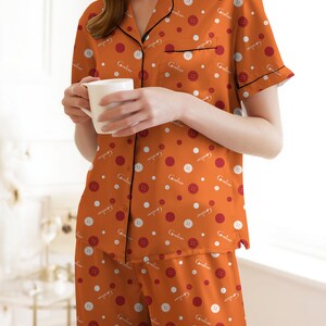 May include: Orange pajama set with a button and polka dot pattern. The short-sleeved top has black trim and a front pocket. The matching pants are a wrap style. The pajamas are made of a soft material.