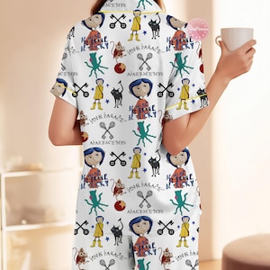 May include: White pajama set with a repeating pattern featuring cartoon characters, cats, keys, and text. The short-sleeved top and pants have yellow trim. The text includes "Pink Palace" and "Be Brave Be Tricky".