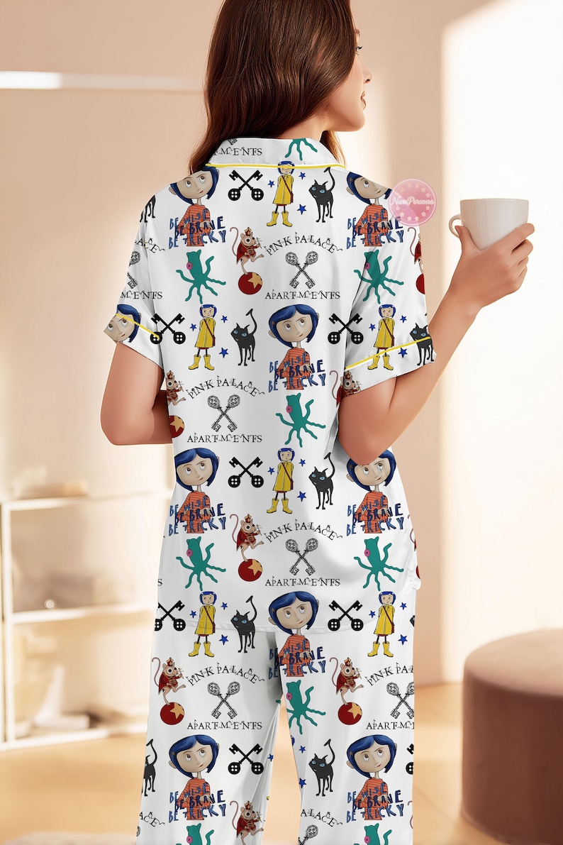 May include: White pajama set with a repeating pattern featuring cartoon characters, cats, keys, and text. The short-sleeved top and pants have yellow trim. The text includes "Pink Palace" and "Be Brave Be Tricky".