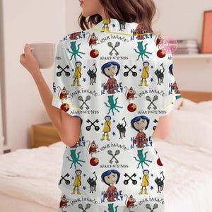 May include: White pajama set with a colorful pattern featuring cartoon characters, text, and various symbols. The short-sleeved top and shorts have yellow trim. The text includes "PINK PALACE", "BE BRAVE", and "APARTMENTS".