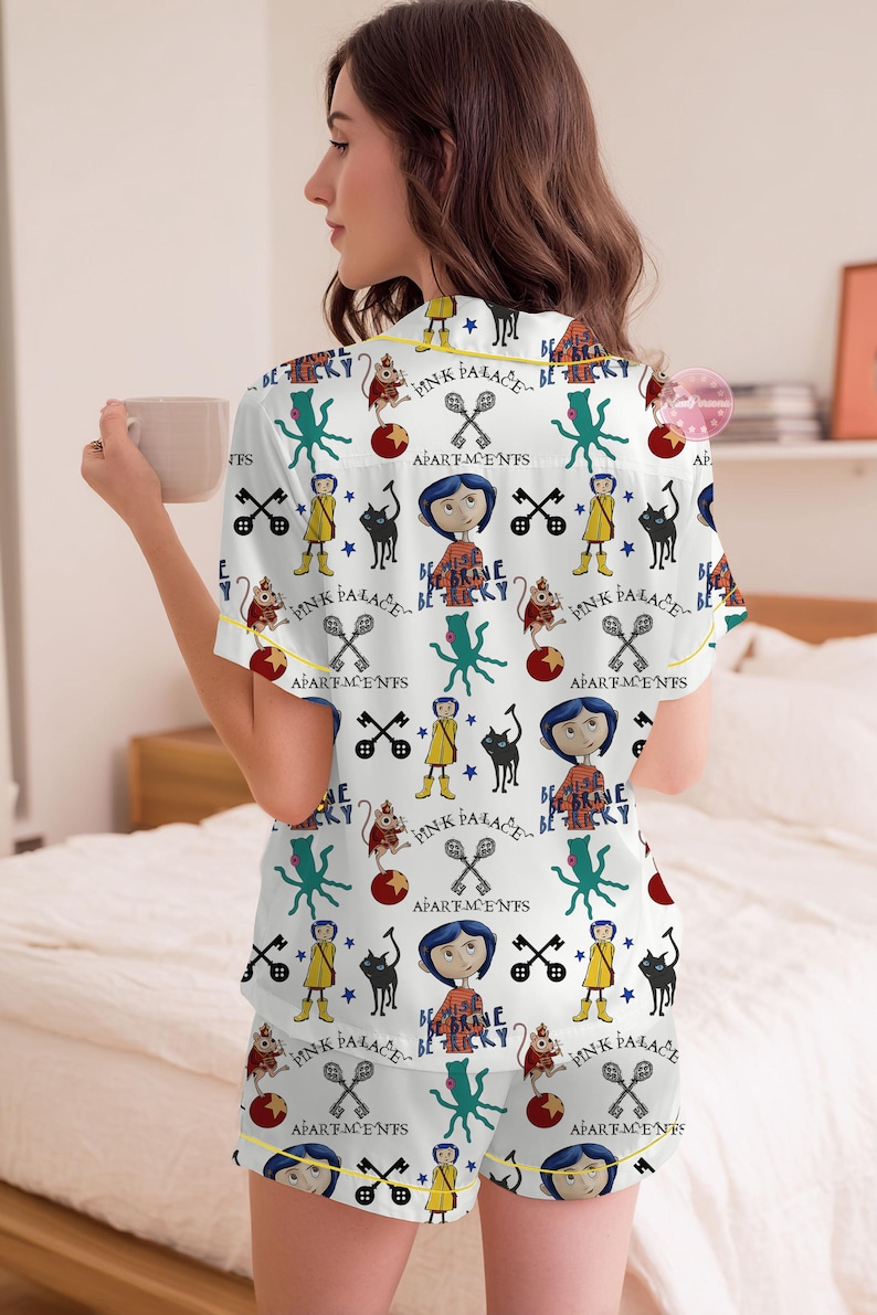 May include: White pajama set with a colorful pattern featuring cartoon characters, text, and various symbols. The short-sleeved top and shorts have yellow trim. The text includes "PINK PALACE", "BE BRAVE", and "APARTMENTS".