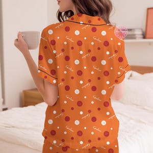 May include: Orange pajama set with a button and polka dot pattern. The short-sleeved top and shorts feature white buttons, red dots, and the name "Caroline" in a script font. Black trim outlines the edges of the set.