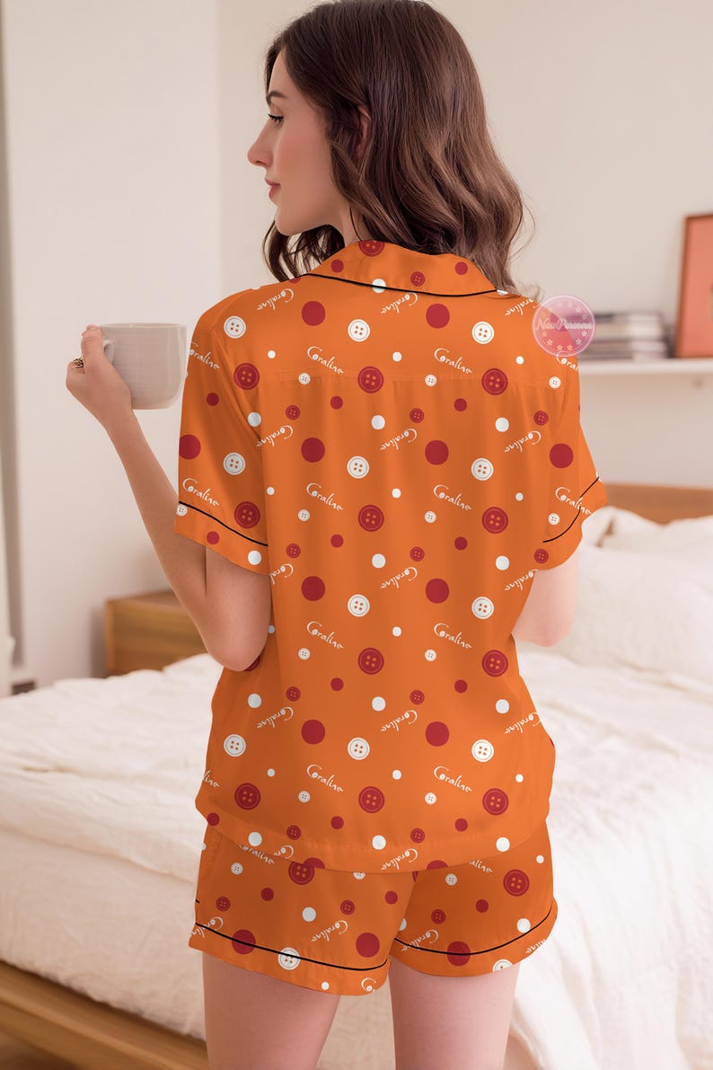 May include: Orange pajama set with a button and polka dot pattern. The short-sleeved top and shorts feature white buttons, red dots, and the name "Caroline" in a script font. Black trim outlines the edges of the set.