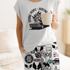 May include: White pajama set with a graphic print. The top features the text "SAVING PEOPLE HUNTING" with a car and a sun symbol. The pajama pants have various Supernatural-themed graphics and text. The person is holding a beige mug.