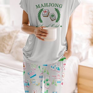 Może przedstawiać: Jasnoszary zestaw piżamowy z topem z kr&oacute;tkim rękawem i pasującymi spodniami. Top ma zielony napis "Mahjong Social Club". Spodnie są wzorzyste z kolorowymi kafelkami Mahjonga i ilustracjami związanymi z grą. Osoba trzyma biały kubek.