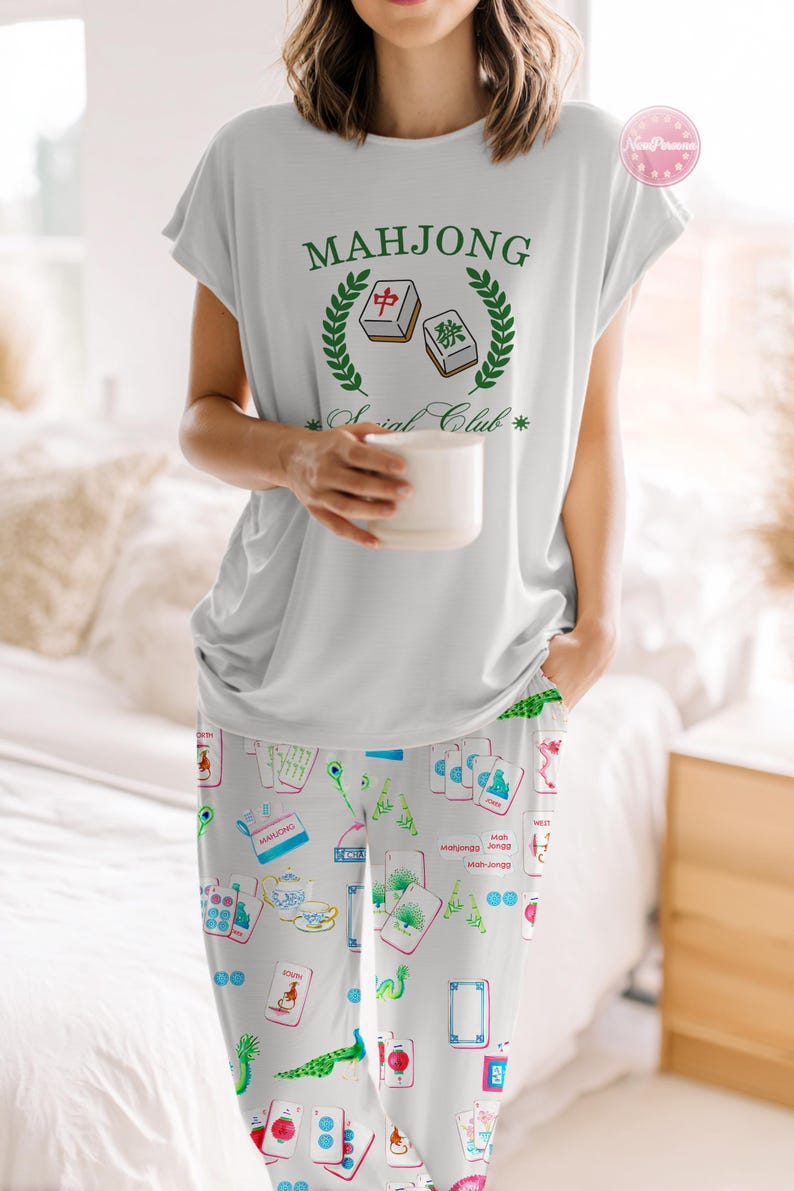 Może przedstawiać: Jasnoszary zestaw piżamowy z topem z kr&oacute;tkim rękawem i pasującymi spodniami. Top ma zielony napis "Mahjong Social Club". Spodnie są wzorzyste z kolorowymi kafelkami Mahjonga i ilustracjami związanymi z grą. Osoba trzyma biały kubek.