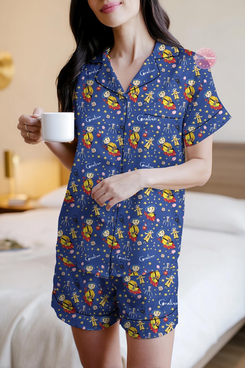 Peut inclure: Ensemble de pyjama bleu marine avec un motif r&eacute;p&eacute;t&eacute; d'un personnage de dessin anim&eacute;, d'&eacute;toiles et du mot "Coraline". Le haut &agrave; manches courtes et le short ont une bordure noire. Une tasse blanche est tenue.