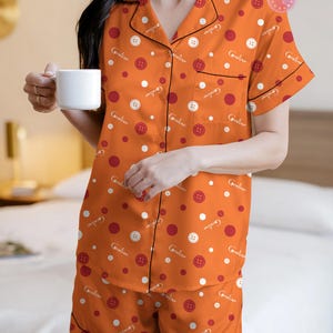 May include: Orange pajama set with white and red polka dots and button graphics. The short-sleeved top and shorts have black trim and the word "Coraline" printed on them. The person is holding a white mug.