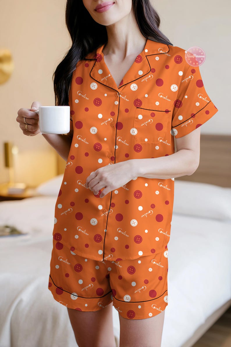 May include: Orange pajama set with white and red polka dots and button graphics. The short-sleeved top and shorts have black trim and the word "Coraline" printed on them. The person is holding a white mug.