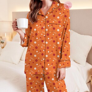 May include: Orange pajama set with a button and dot pattern. The long-sleeved top has black trim and a breast pocket. The matching pants complete the set. The pajamas have the word "Caroline" printed on them. A white mug is held in the hand.
