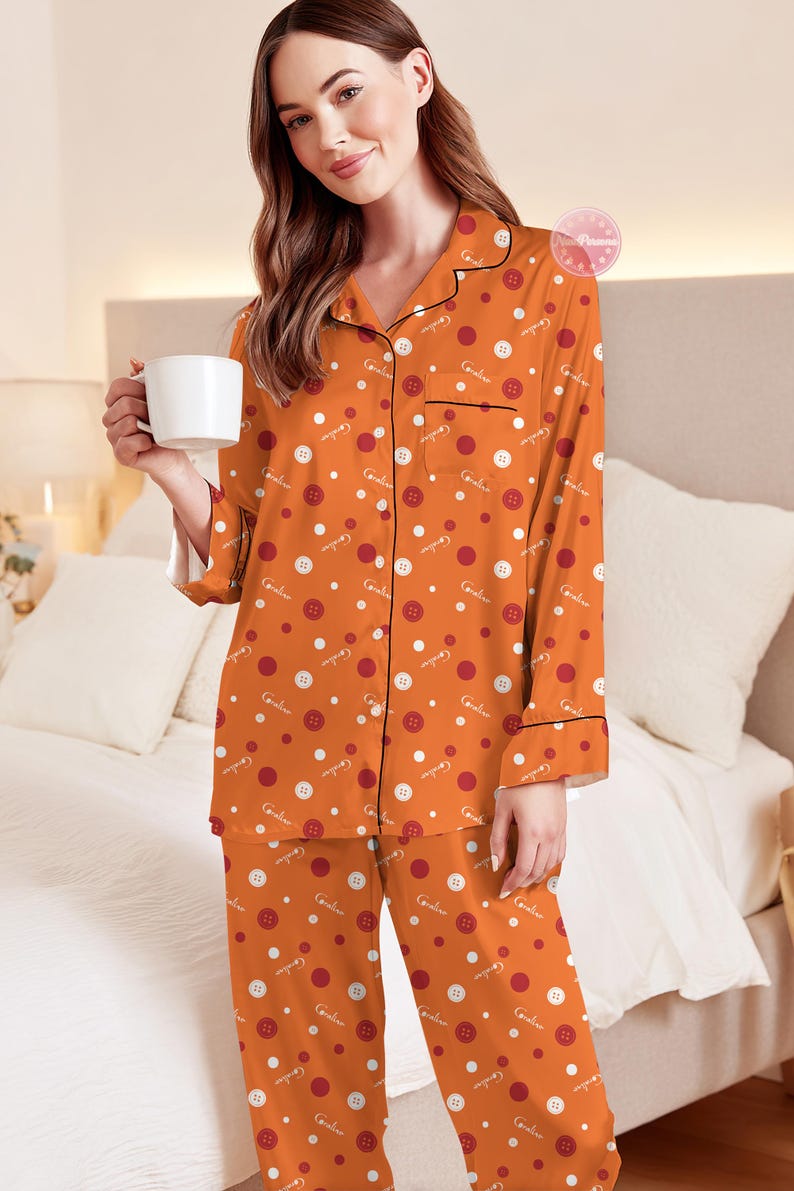 May include: Orange pajama set with a button and dot pattern. The long-sleeved top has black trim and a breast pocket. The matching pants complete the set. The pajamas have the word "Caroline" printed on them. A white mug is held in the hand.