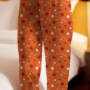 May include: Orange pajama pants with a pattern of red buttons, white buttons, and red circles. The word "Coraline" is written in white script throughout the design. The pants are made of a soft fabric.