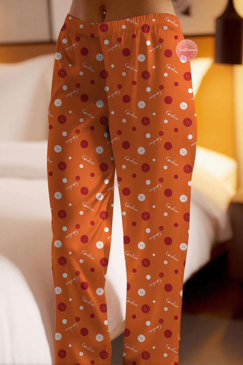 May include: Orange pajama pants with a pattern of red buttons, white buttons, and red circles. The word "Coraline" is written in white script throughout the design. The pants are made of a soft fabric.
