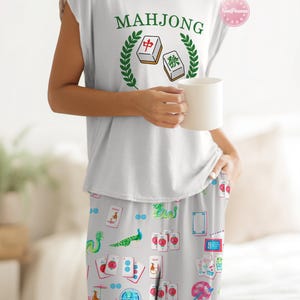 Może przedstawiać: Biały zestaw piżamowy. Bezrękawnik z napisem "MAHJONG" i grafiką płytek Mahjonga. Pasujące spodnie od piżamy mają jasnoszare tło z kolorowymi ilustracjami z motywem Mahjonga. W dłoni trzymany jest biały kubek.