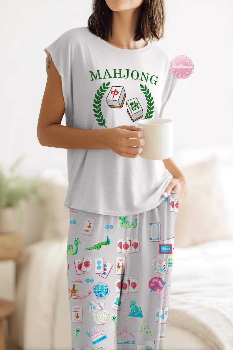 Może przedstawiać: Biały zestaw piżamowy. Bezrękawnik z napisem "MAHJONG" i grafiką płytek Mahjonga. Pasujące spodnie od piżamy mają jasnoszare tło z kolorowymi ilustracjami z motywem Mahjonga. W dłoni trzymany jest biały kubek.