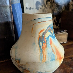 May include: A ceramic vase with a cream-colored base and a marbled design in shades of blue, orange, and yellow. The vase has a rounded body and a slightly flared neck, sitting on a wooden surface.