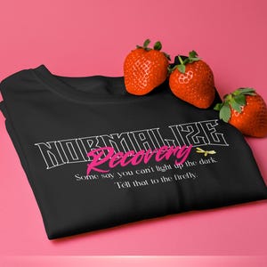 May include: Black t-shirt with the words "NORMAL LIFE Recovery" in white and pink, with a dragonfly graphic. The shirt has the quote "Some say you can't light up the dark. Tell that to the firefly." Three strawberries rest on the shirt.