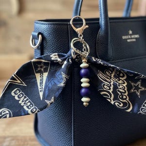 May include: A navy blue handbag with a decorative keychain. The keychain features a scarf with "Dallas Cowboys" text and star graphics, along with a beaded charm in white and purple. The bag has a textured finish.