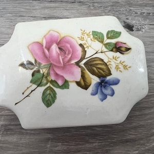 May include: A rectangular, white ceramic trinket box with a floral design. The lid features a pink rose, a blue flower, and green leaves. The box has a slightly curved shape and a glossy finish.
