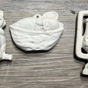 May include: Three unpainted ceramic ornaments. One is a raccoon holding a sack, another is a baby in a basket, and the third is a lantern with a bear. Each has a hole for hanging.