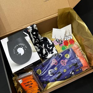 May include: An open cardboard box filled with Halloween-themed items. Includes a black and white patterned scarf, a candle with the text "I'll put a smell on you", a pair of purple socks with skulls, and earrings with ghost and Scream designs.