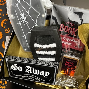 May include: A Halloween-themed gift box filled with various items. Includes a coffin-shaped box with a spiderweb design, a notebook, an eye mask with "Go Away" text, and a candle. The box is lined with gold tissue paper.