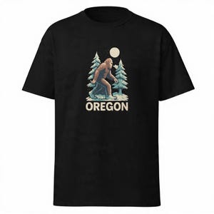 Puede incluir: Camiseta negra con un diseño de Bigfoot caminando en un bosque nevado bajo la luna llena. La palabra "OREGON" está impresa en blanco debajo de la ilustración. El diseño está centrado en la parte delantera.