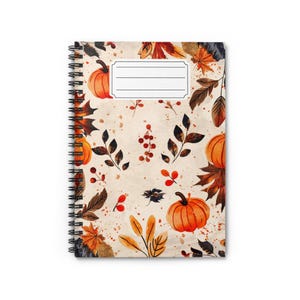 May include: A spiral-bound notebook featuring a cream-colored cover with a repeating pattern of orange pumpkins, brown and red autumn leaves, and small red berries. A white rectangular label with four horizontal lines is centered at the top.