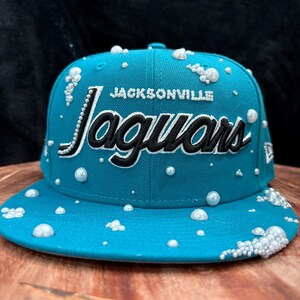 May include: A teal baseball cap with the text "Jacksonville Jaguars" in black and white. The cap is adorned with white pearl-like embellishments. The hat has a curved brim and a structured crown.