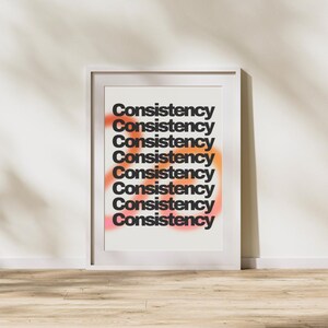 Puede incluir: Un póster enmarcado con la palabra "Consistency" repetida en negro, apilada verticalmente. El fondo presenta un degradado de colores naranja y rosa. La obra de arte se exhibe contra una pared blanca, con sombras.