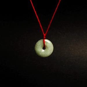 Pode incluir: Um colar com pingente de donut de jade com um cordão vermelho sobre um fundo preto. O pingente é de cor verde claro com um buraco no centro. O cordão vermelho é fino e parece ser de seda.