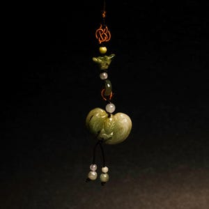 Puede incluir: Un colgante de jade con un diseño en forma de corazón, con una mezcla de cuentas verdes y blancas. El colgante está suspendido de un cordón negro y tiene una apariencia delicada y artesanal. El diseño general es orgánico y natural.