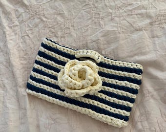 May include: A handmade, striped clutch in navy blue and cream, adorned with a cream-colored crocheted flower. The clutch features horizontal stripes and a rectangular shape, perfect for carrying essentials. The design is simple yet elegant.