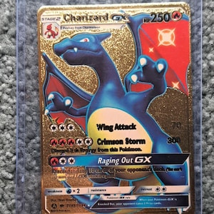 May include: A gold Charizard GX Pokémon trading card. The card features a blue and black dragon with red accents. The card has the text "Wing Attack", "Crimson Storm", and "Raging Out GX". The card is in a protective sleeve.