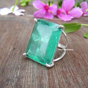 Pode incluir: Um anel de prata com uma grande pedra preciosa retangular verde esmeralda. O anel tem um design simples e moderno com um acabamento polido. A pedra preciosa é o ponto focal, refletindo um tom verde vibrante. O anel está sobre uma superfície de madeira.