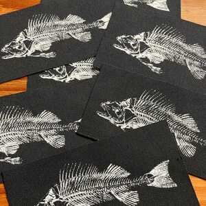 May include: A collection of rectangular black fabric patches, each featuring a white screen-printed graphic of a fish skeleton. The skeletal design displays the detailed bone structure of the fish, including the skull, spine, and fins, in high contrast.