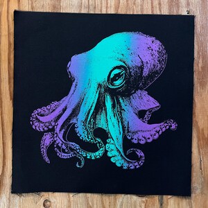 May include: A square black fabric patch featuring a detailed screen-printed illustration of an octopus. The octopus design displays a vibrant gradient color transition, shifting from bright cyan on the left to deep purple on the right side.