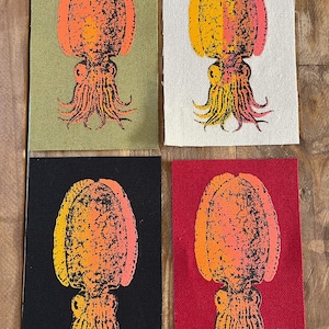 Op de afbeelding: Vier rechthoekige stoffen patches met een gezeefdrukt inktvisontwerp. De inktvis is oranje en geel met zwarte details. Elke patch heeft een andere achtergrondkleur: olijfgroen, wit, zwart en rood. De patches zijn gerangschikt in een 2x2 raster.
