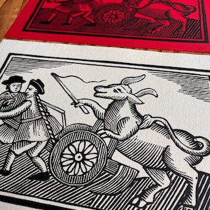 Op de afbeelding: Twee rechthoekige prints met een houtsnede-illustratie in zwarte inkt van een persoon met een hoed die een tweewielige kar bestuurt, getrokken door een gehoornd wezen met een vlag, afgebeeld op een witte en een rode achtergrond.