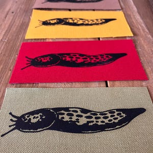 May include: A selection of fabric patches with a black slug design. The patches are displayed in a range of colors: white, brown, yellow, red, and olive green. The slug illustration is a key feature on each patch.