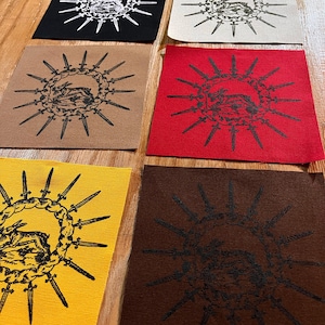 Puede incluir: Seis parches de tela cuadrados con un gráfico central de un conejo rodeado por un círculo de espadas. Los parches vienen en negro, blanco roto, beige, rojo, amarillo y marrón, cada uno con la misma ilustración detallada en tinta.