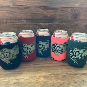 May include: A collection of five beverage can coolers in various colors: black, red, navy, coral, and green. Each cooler features a green frog graphic. Cans with red and white labels are visible inside the coolers. The coolers are arranged on a wooden surface.