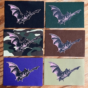 May include: Six rectangular fabric patches featuring a printed graphic of a flying bat. The patches come in black, dark green, camouflage, brown, purple, and olive green. Each patch displays the same bat illustration in a light pinkish hue.
